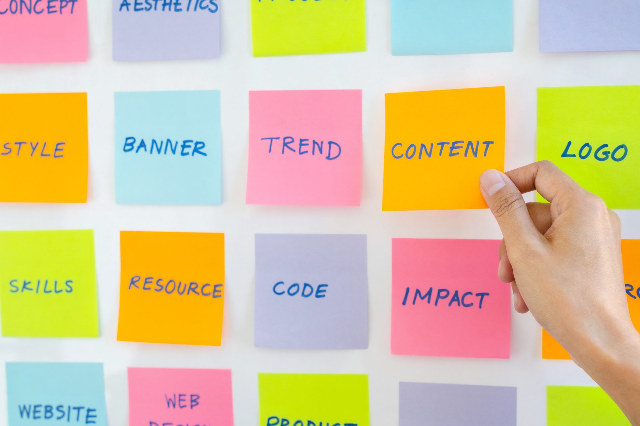 UX UI applications designer working for smart phone interface. Hand holding an orange sticky note labeled "CONTENT" among colorful sticky notes with words like "TREND," "BANNER," "IMPACT," and "LOGO," representing creative brainstorming and storytelling concepts for a photographer and visual storyteller.
