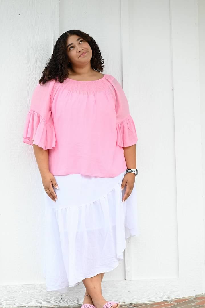 SflyProxyVersion 1; Woman with curly hair wearing a pink blouse and white skirt standing thoughtfully against a white wooden wall, embodying creative storytelling and soulful fine art photography.